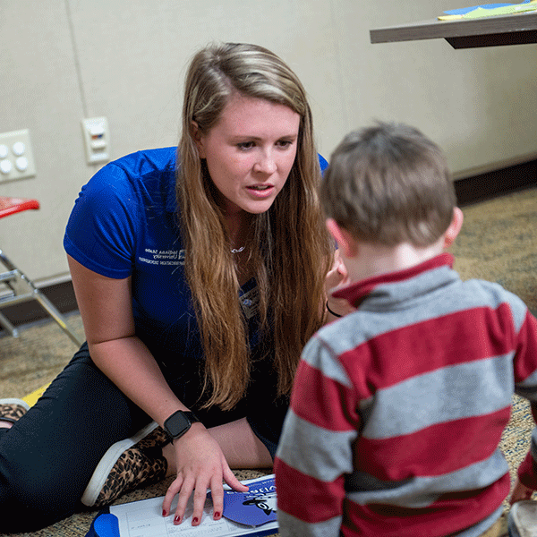 A white female student with long blond hair is sitting on the ground with a toddler boy with short brown hair. The female student wears a blue Indiana State University T-shirt, blue jeans, and cheetah-print shoes. The toddler wears a red-and-grey long-sleeved shirt.