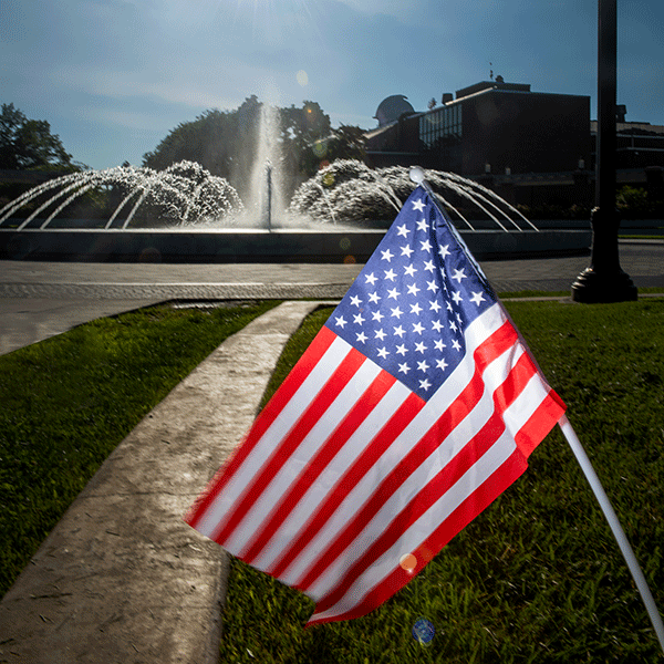 An American flag with white stars on a blue background, and red and white stripes in front of a fountain with one spout in the center and several small spouts around the center in a circular manner.